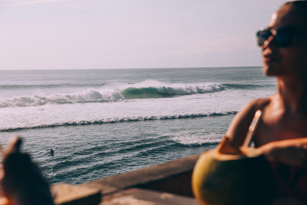 california beach and waves surfing and girl sitting enjoying paradise sunrise with sunglasses and drinking from coconut, thank the wavemaker clothing brand surf brand